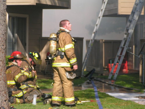 Fireman on the job looking at burning house and other is sitting.