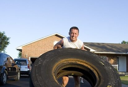 A man using DIY gym equipment to save money by lifting a large tire in front of a house.