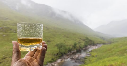 A person holding a glass of whisky in front of a mountain, showcasing the Art of Manliness.
