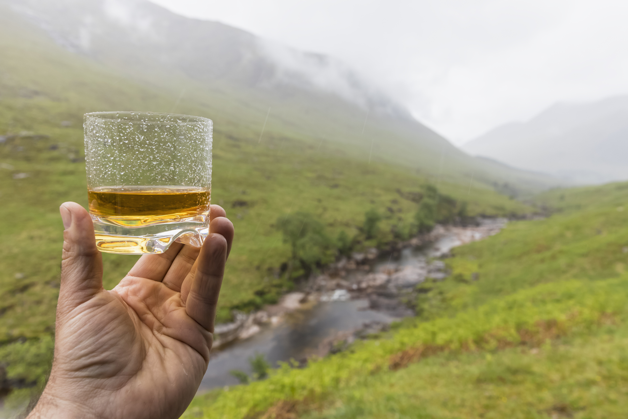A person holding a glass of whisky in front of a mountain, showcasing the Art of Manliness.