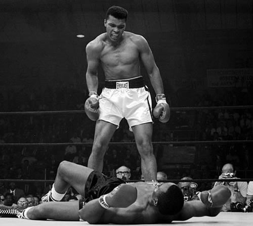 Muhammad Ali stands over Sonny Liston in boxing ring.