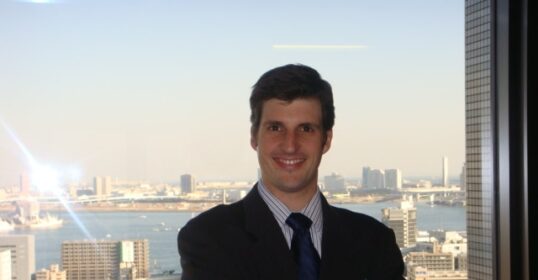 A man in a suit standing in front of a city view, appearing as an interpreter or translator.