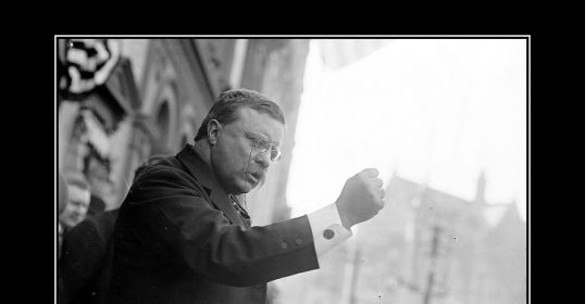A man in a suit is holding a sign that says vigor, inspired by Theodore Roosevelt.