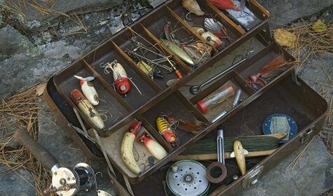 A man with a fishing tackle box carefully arranges the basics of fishing lures in a suitcase on the ground.