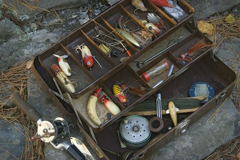 A man with a fishing tackle box carefully arranges the basics of fishing lures in a suitcase on the ground.