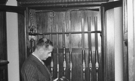 A man in a suit standing in front of a gun cabinet, showcasing his home defense arsenal.