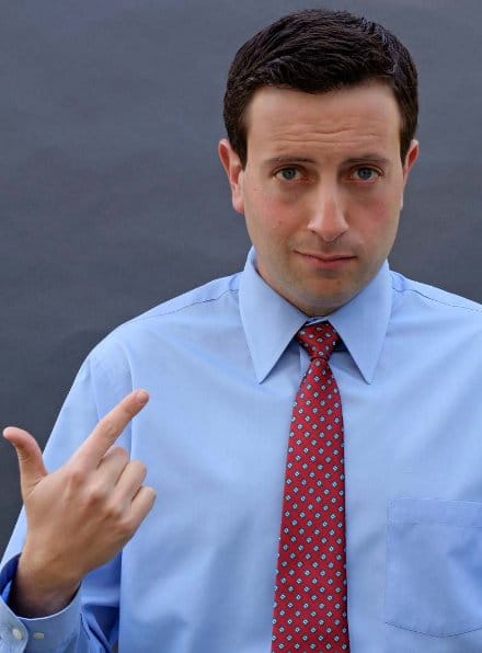 An actor in a blue shirt and tie making a fist gesture in his job.