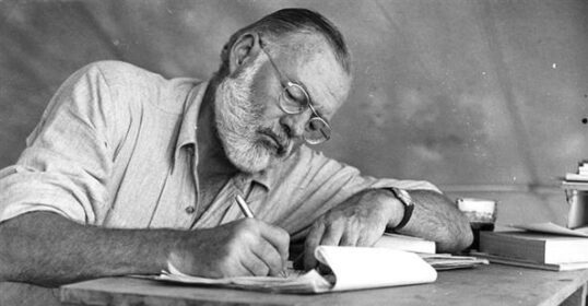Ernest Hemingway, a man sitting at a table writing a book with motivational posters in the background.