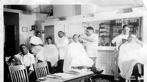 A black and white photo of men in a barber shop, getting their hair cut by a pick.