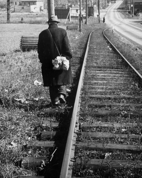 A hobo walking down the railroad tracks in an old black and white photo.