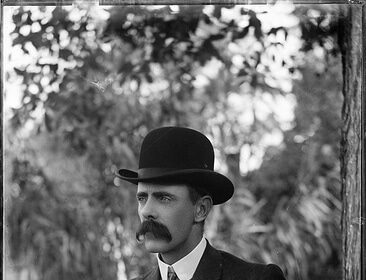 A man in a suit and hat, with a mustache, sitting on a bench.
