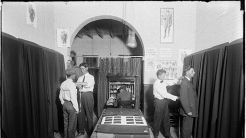 A group of men standing in front of a room with a curtain, discussing clothing alterations.