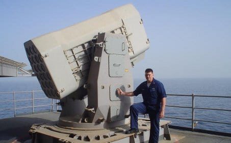 A Navy Sailor standing next to a missile on the deck of a ship.