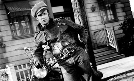 A man in a leather jacket leaning on a motorcycle, offering breaking in tips for his new jacket.
