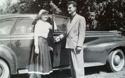 A man and woman standing next to an old car in a chivalrous pose, embodying the Art of Manliness.
