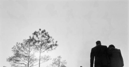 A black and white photograph of a crowd of people walking in the grass, with a man following funeral etiquette.