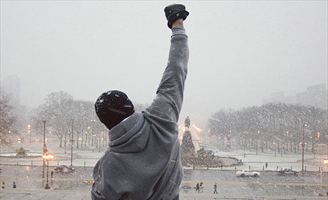 A man in a hoodie raises his fist in the snow, as if ready to engage in a boxing match.