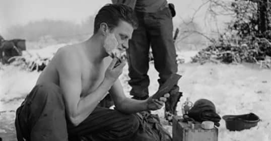 A soldier shaving outdoors in a snowy environment using cold water and a hand mirror.