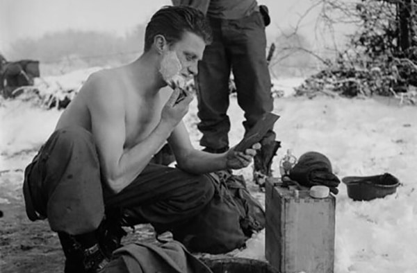 A soldier shaving outdoors in a snowy environment using cold water and a hand mirror.