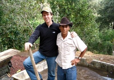 Two men standing next to a well in a wooded area, discussing their job as coffee buyers.