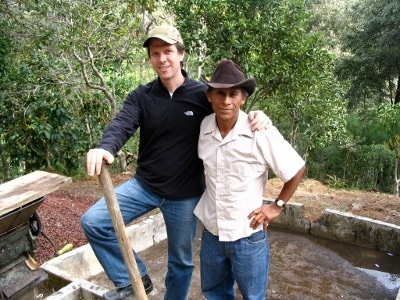 Two men standing next to a well in a wooded area, discussing their job as coffee buyers.