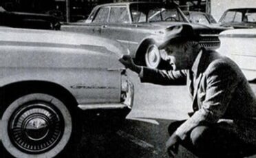 A man kneeling down next to a parked car, contemplating whether to buy the used car.