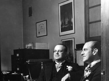 Two best men in suits discussing their duties in a room.