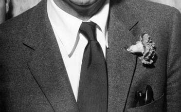 A man wearing a suit with a tie and a boutonniere pinned to the left lapel, partial view of his head, upper torso, and jacket pocket visible.