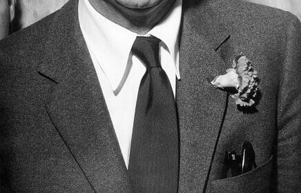 A man wearing a suit with a tie and a boutonniere pinned to the left lapel, partial view of his head, upper torso, and jacket pocket visible.