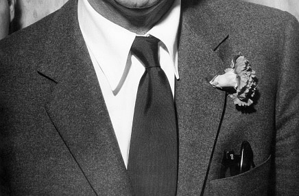 A man wearing a suit with a tie and a boutonniere pinned to the left lapel, partial view of his head, upper torso, and jacket pocket visible.
