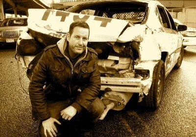 A stuntman kneeling next to a wrecked car.