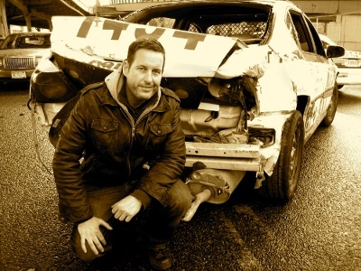 A stuntman kneeling next to a wrecked car.