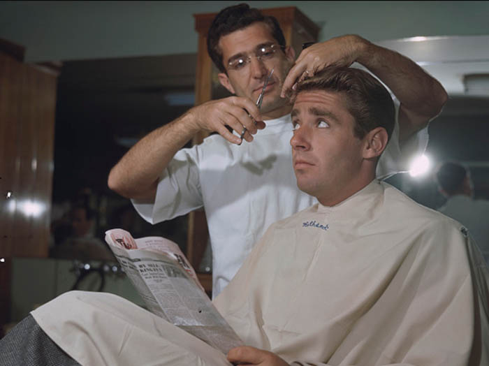 The man is getting a haircut while talking to the barber in a barbershop.