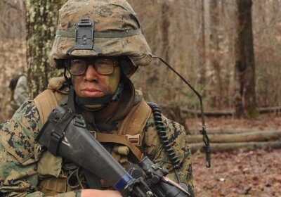 A female Marine Corps Officer crouching in the woods with a rifle, executing her job with precision.