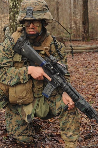 A female Marine Corps Officer crouching in the woods with a rifle, executing her job with precision.