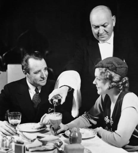 A waiter elegantly pours a glass of wine to a man and woman on their dinner date.