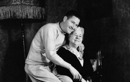A black and white photo of a man and woman sitting on a chair, depicting a mother and man in a loving relationship.