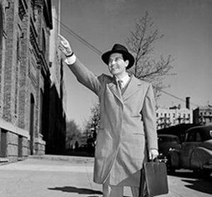 A man in a suit and hat hailing a taxi cab on a sidewalk.