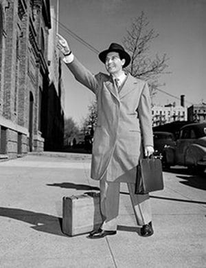 A man in a suit and hat hailing a taxi cab on a sidewalk.