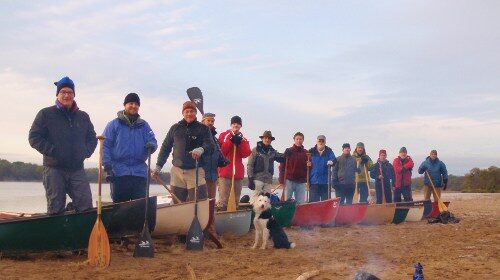 A successful group of people plan a canoe trip, standing next to canoes on the beach.