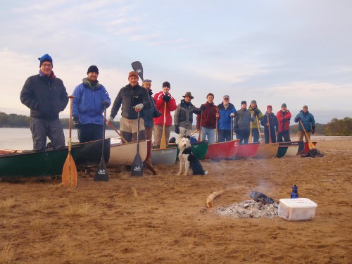 A successful group of people plan a canoe trip, standing next to canoes on the beach.