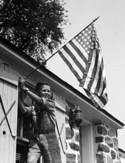 Verouderde jongen die de Amerikaanse vlag buiten het raam van zijn huis hangt. 