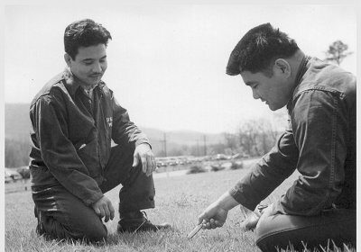 Two men playing Mumbley Peg, kneeling in the grass and looking at a piece of paper.