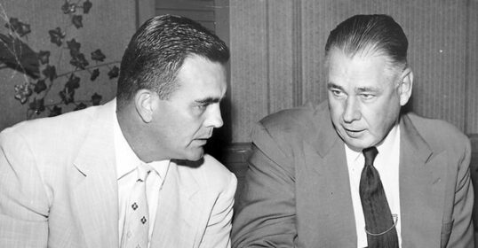 Two men, John and Michael, sitting at a table. John tries to remember something but can't.