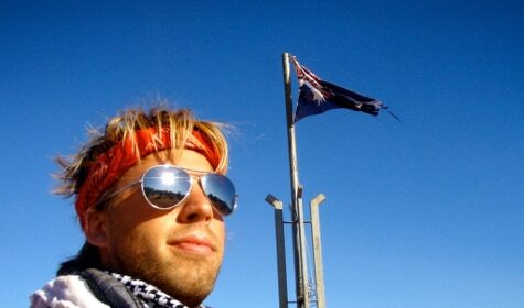 A Motorcycle Expedition Guide confidently poses in front of an Australian flag, sporting sunglasses and a trendy scarf.