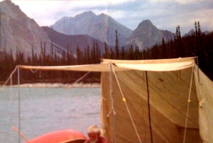 An unconventional man in a tent next to a canoe on a lake.