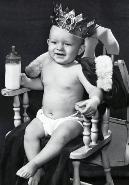 Vintage infant baby sitting on the chair wearing king crown and feeder in the hand.