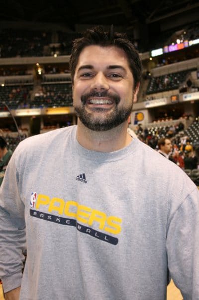 A man smiling in front of a basketball court, likely an NBA Strength and Conditioning Coach.