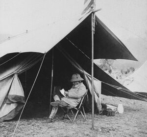 A manly man sitting in front of a tent reading a book.