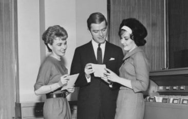 Three women standing in front of an office desk, observing a piece of paper.
